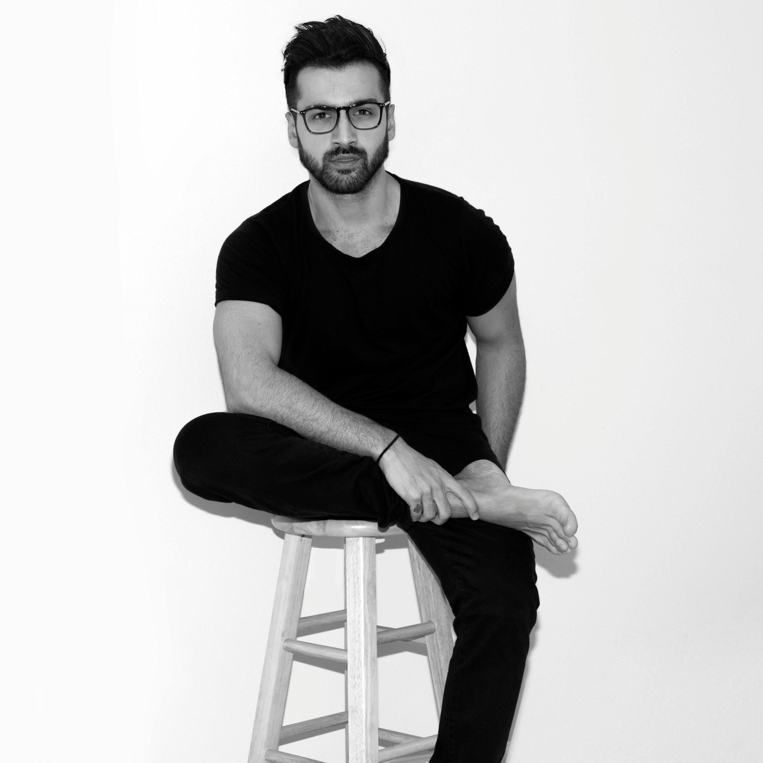 Black and white photo of a man sitting on a stool indoors, looking pensive and fashionable.